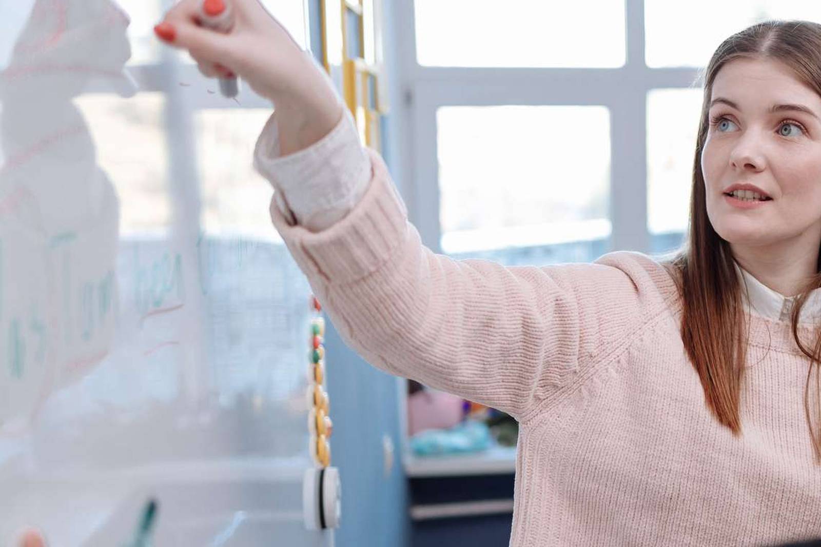 A woman in a pale pink sweater points a marker at a whiteboard while explaining something