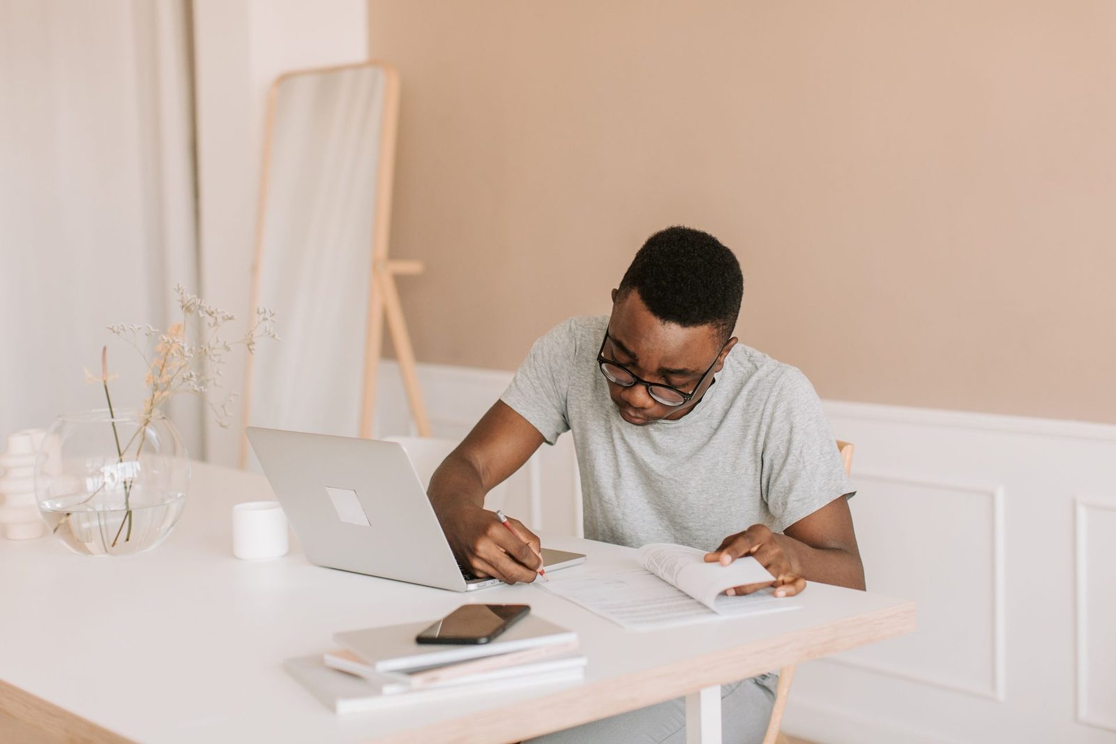 A man wearing glasses writes in a notebook at a home desk beside an open laptop