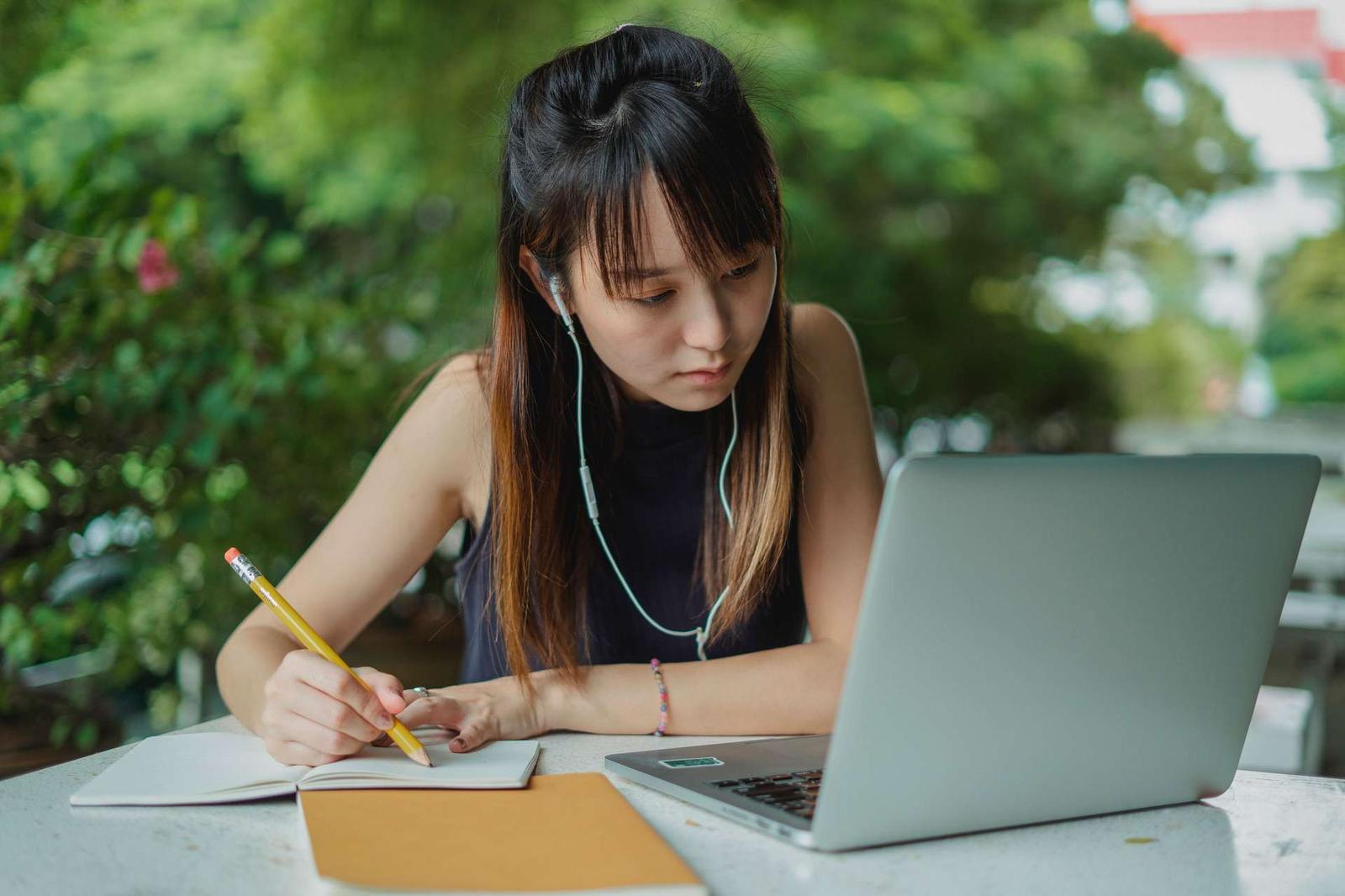 A young woman sits at an outdoor table writing in a notebook with a pencil, a laptop open beside her