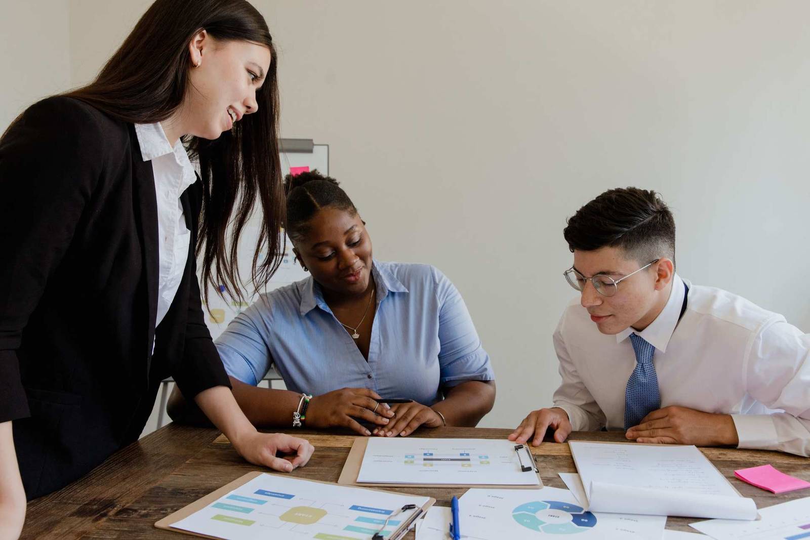 Three colleagues lean over a table covered in printed flowcharts and notes, two seated and one standing, in a collaborative discussion
