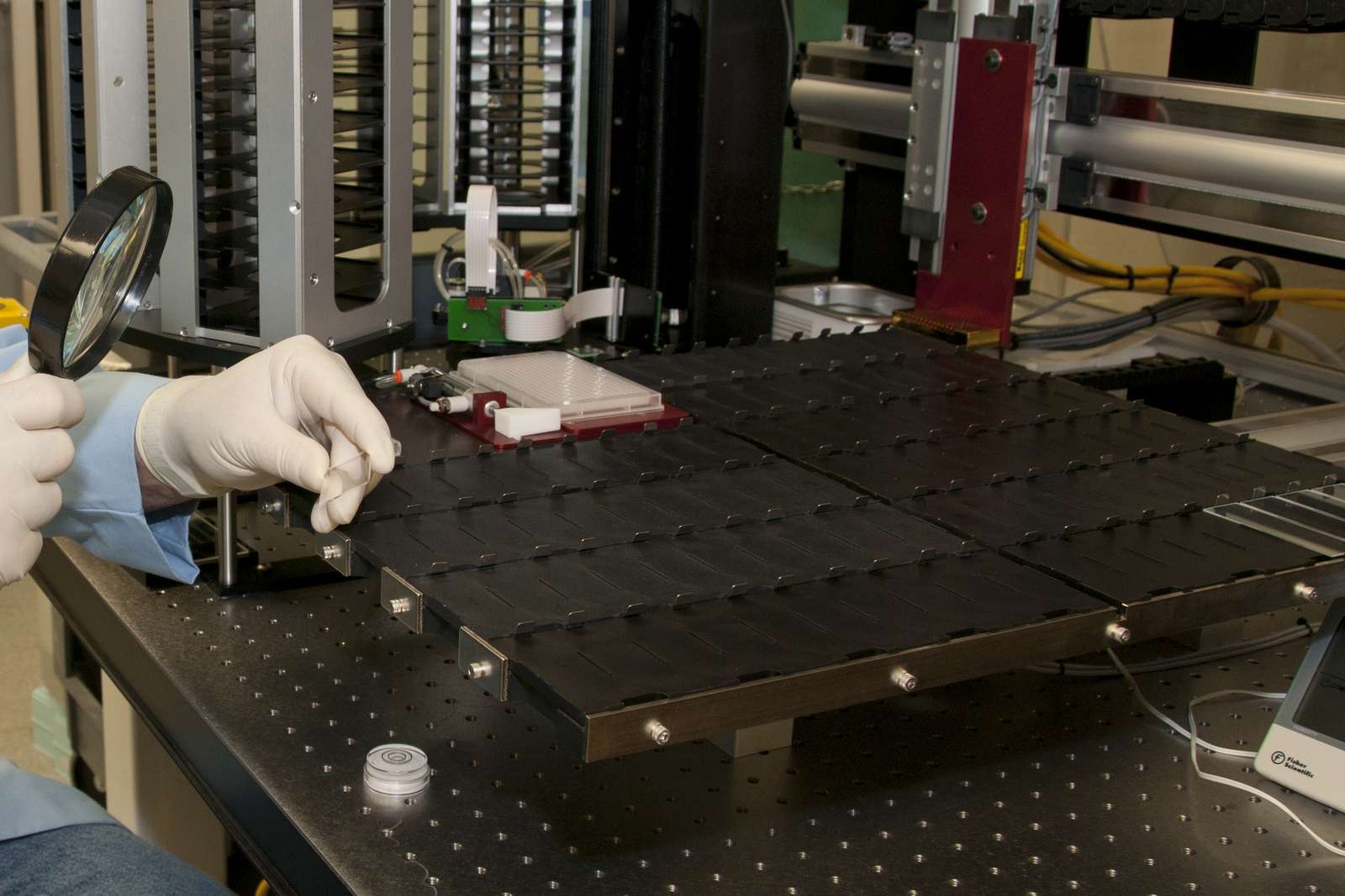 Gloved hands hold a magnifying lens and a glass slide beside a laboratory microarray instrument
