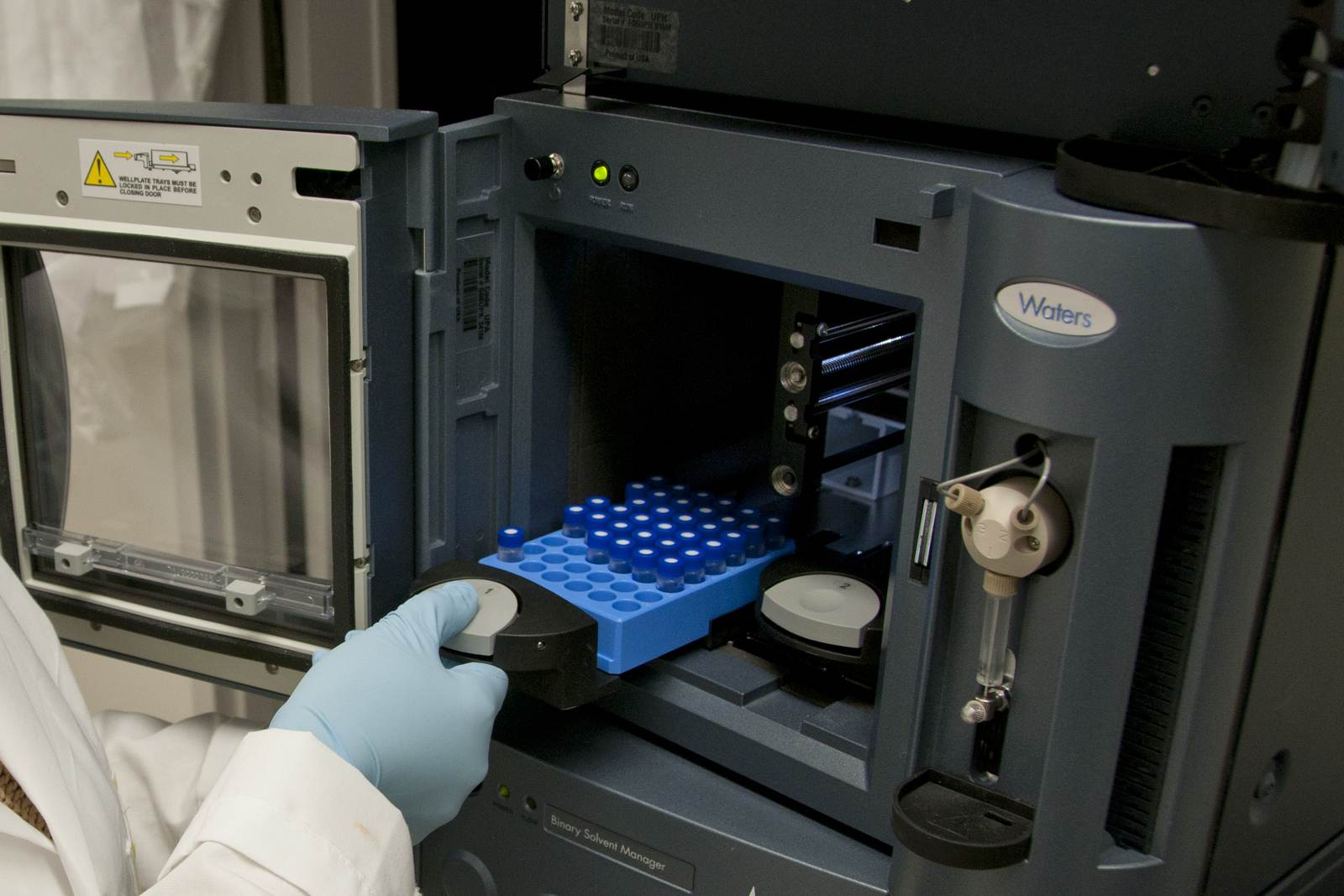 A gloved hand loads a tray of capped sample vials into a laboratory autosampler