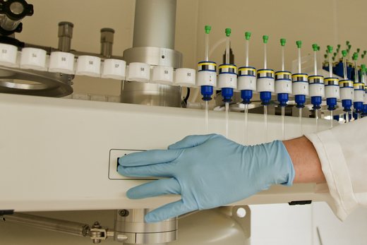 A gloved hand rests on an automated laboratory instrument beside numbered collection positions and rows of capped tubes