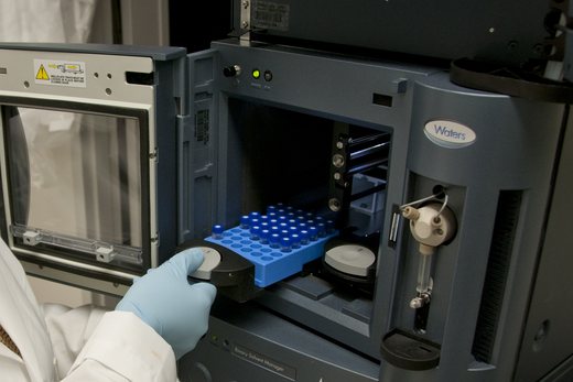 A gloved hand loads a tray of capped sample vials into a laboratory autosampler